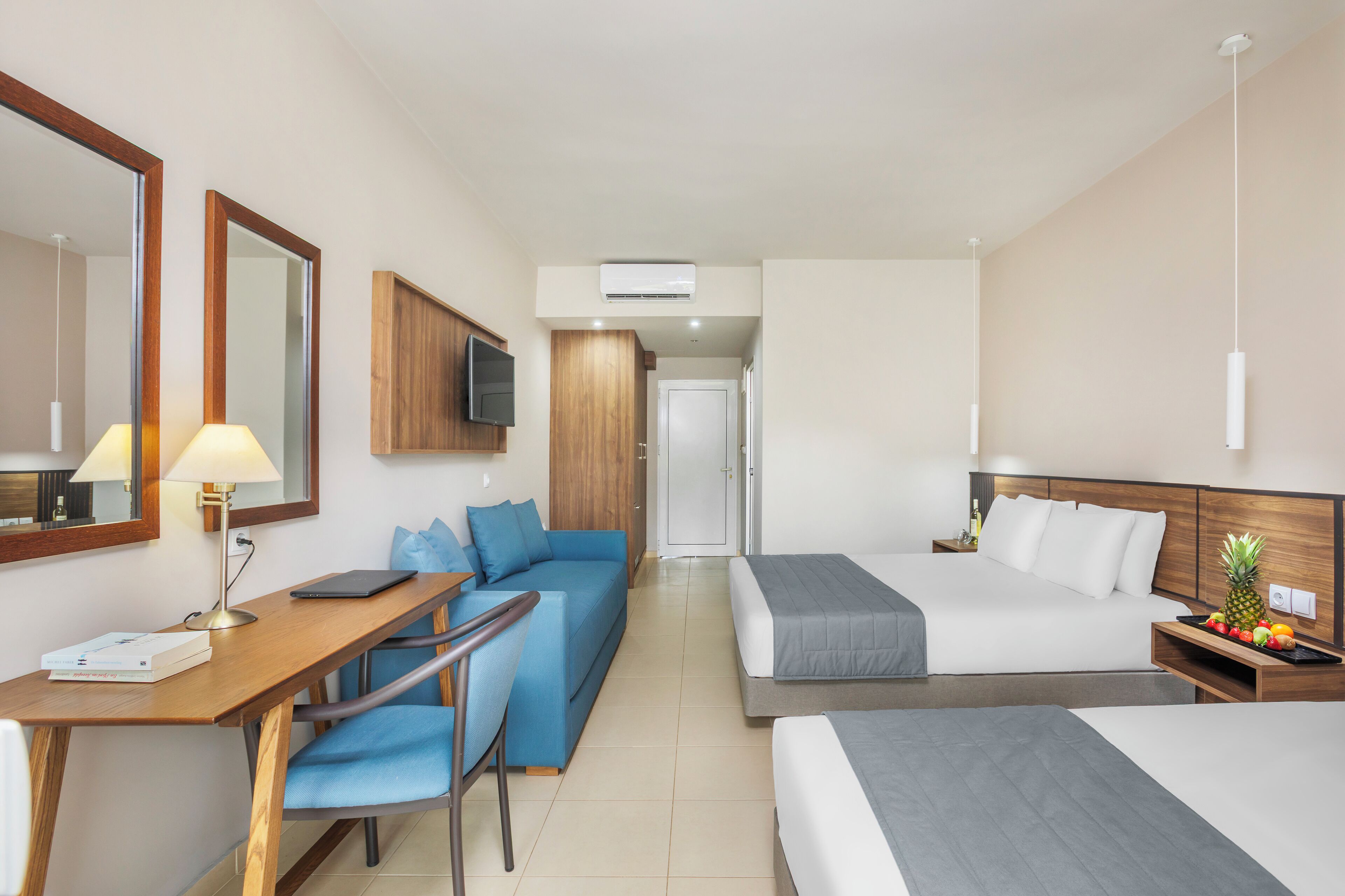 Habitación de hotel con escritorio, sofá azul, dos camas, televisión y una bandeja de fruta.