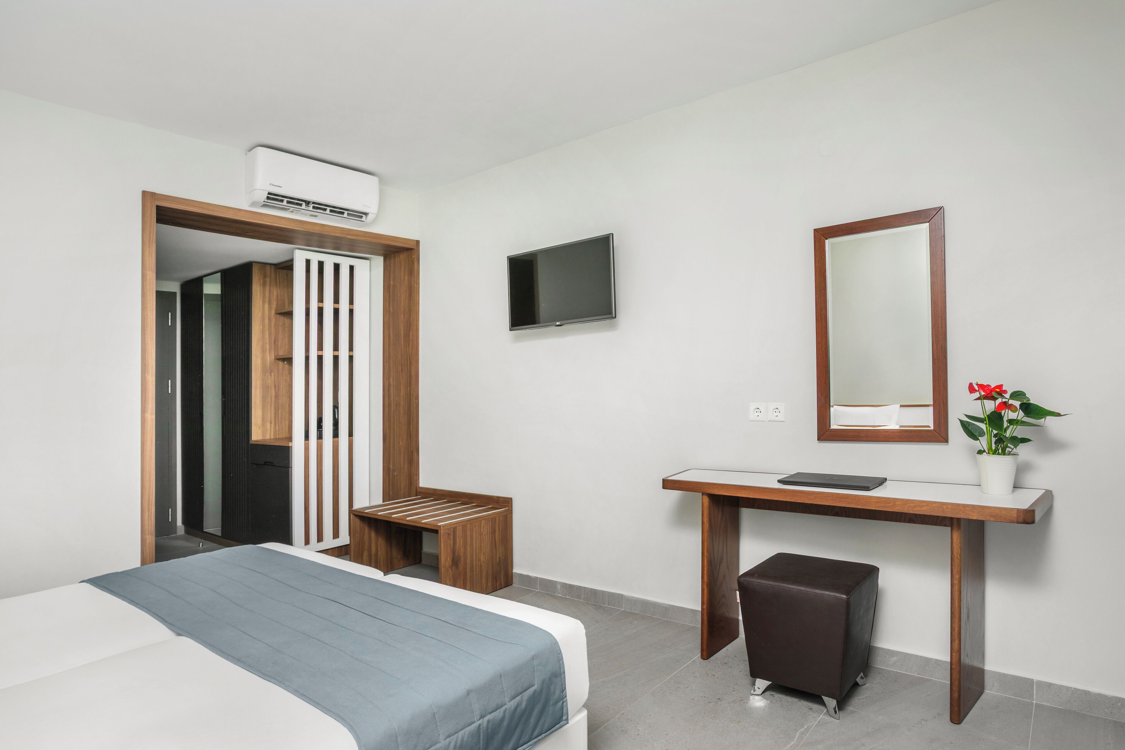 A modern hotel room with a bed, a desk with a mirror, a TV, and an air conditioning unit.