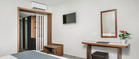 A modern hotel room with a bed, a desk with a mirror, a TV, and an air conditioning unit.