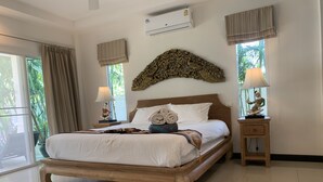A spacious bedroom with a large bed, a wooden headboard, and a decorative wall piece above it. There are two bedside tables with lamps, a window with curtains, and a view of greenery outside. - PACOTTE, nice living villa with private (Rawai)