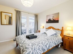 A bedroom with a bed, a nightstand, a lamp, a window with curtains, a painting on the wall, and a view of the outside. - Coastal Bolthole - Great accommodation on the North Norfolk coast. (Kings Lynn)