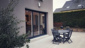 A modern house with a patio area featuring a table and chairs, a sliding glass door, and a well-maintained hedge.
