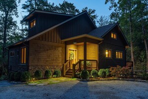 A two-story log cabin with a stone foundation, a front porch with a staircase, and a well-lit entrance.