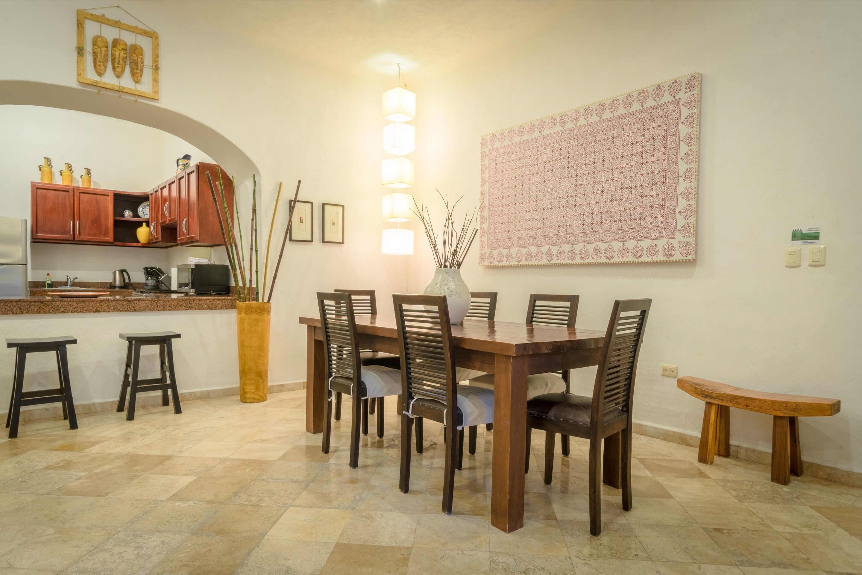 A dining area with a wooden table and chairs, a vase with branches, and a kitchen in the background.