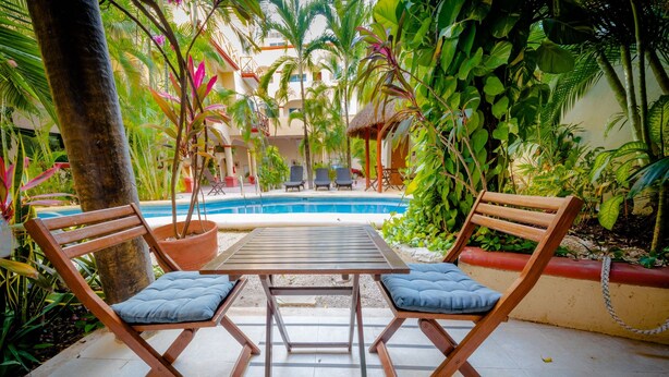 Zona de piscina amb dues cadires de fusta i una taula, envoltada de vegetació tropical exuberanta i una piscina blava cristal·lina.
