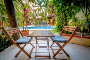 Una zona junto a la piscina con dos sillas de madera y una mesa, rodeada de exuberante vegetación tropical y una piscina azul cristalina.