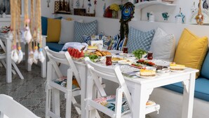 A dining area with white furniture, a table set with food, and a blue sofa with yellow cushions. - Hotel Lime Bozcaada (Bozcaada)