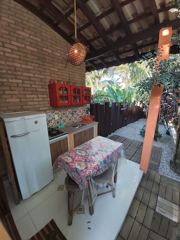 Una cocina con refrigerador, estufa y pileta. Hay una mesa con mantel floral y una silla. La cocina tiene una pared de ladrillo y techo de madera.
