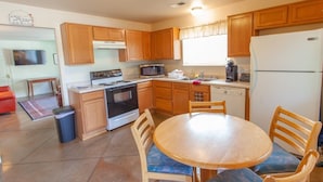 A compact kitchen with wooden cabinets, a stove, microwave, and a refrigerator. There is a dining table with chairs and a trash bin.