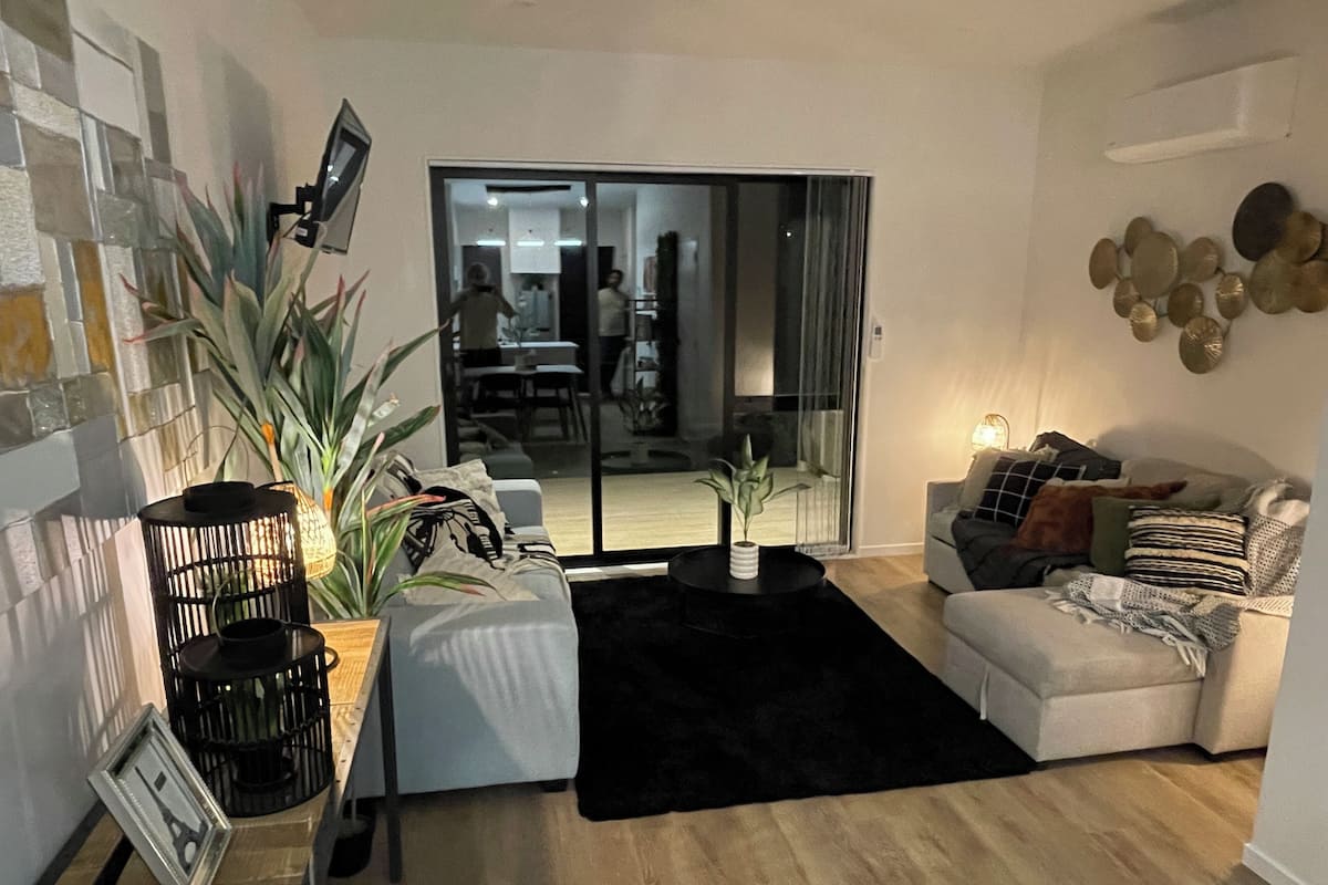 Ett modernt vardagsrum med en soffa, ett soffbord och en stor krukväxt. En skjutdörr i glas leder in till en matplats.