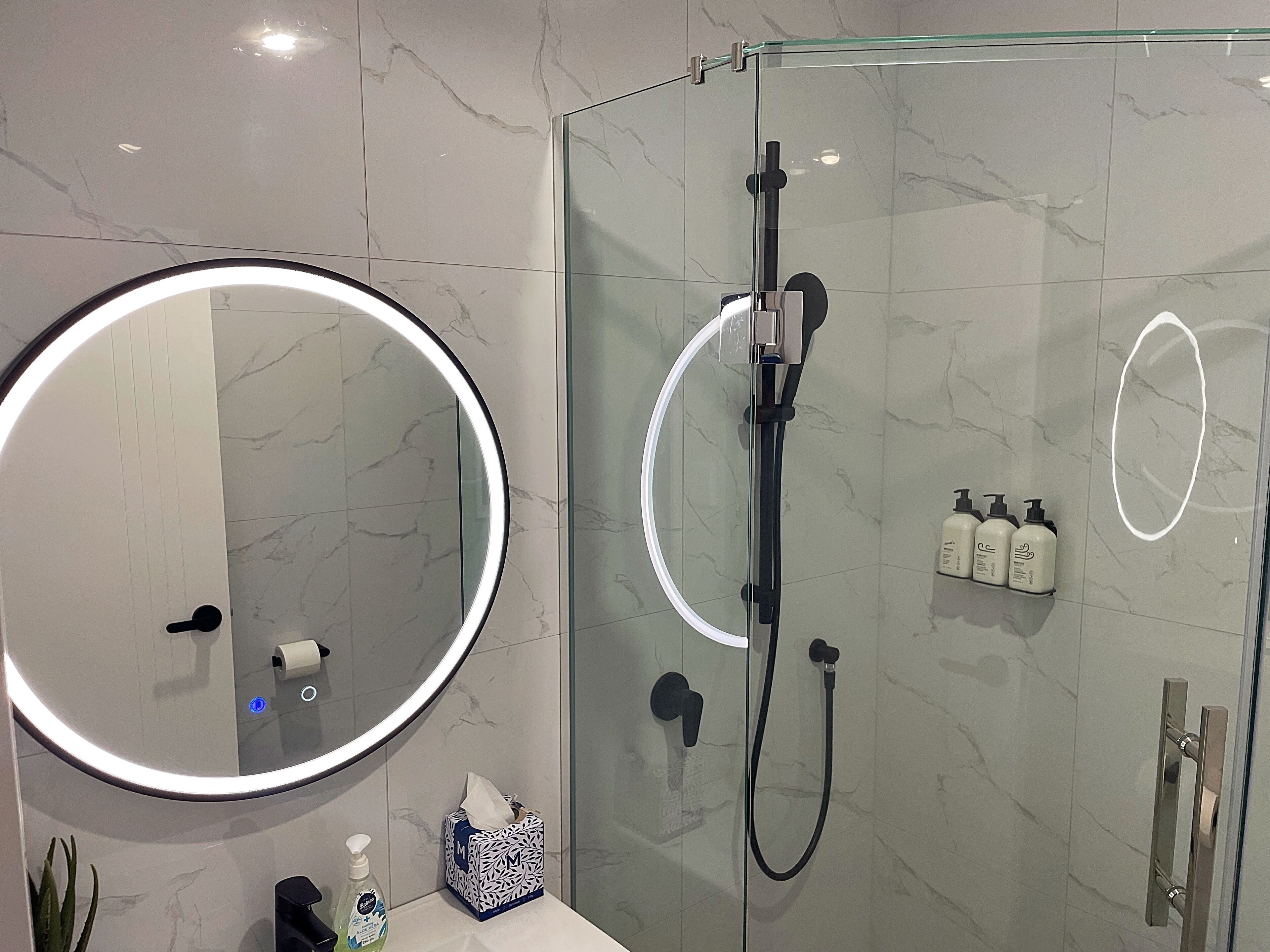 Un baño moderno con ducha envuelta en vidrio, espejo redondo con luz, y artículos de aseo en la encimera.