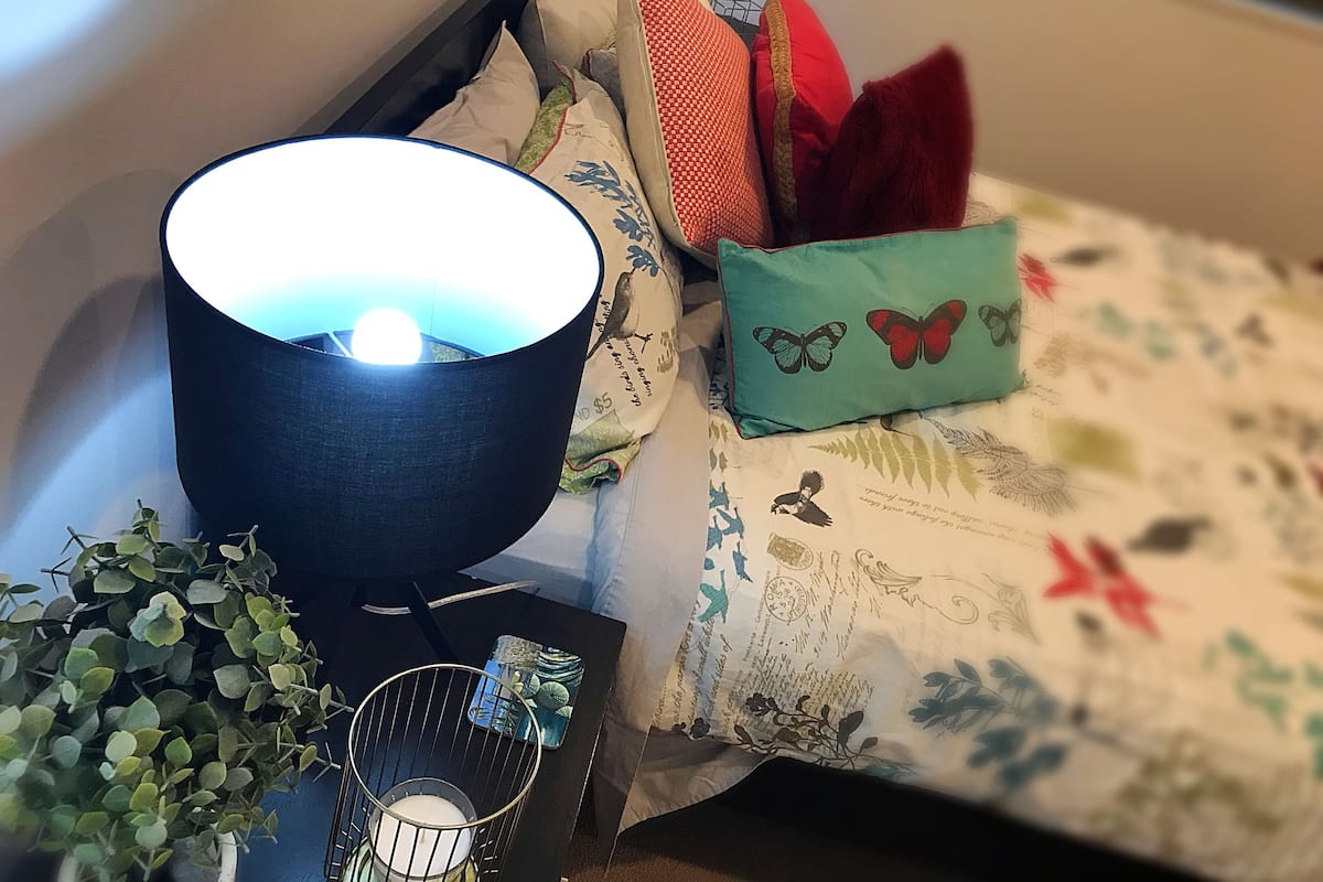 Ett litet bord som står vid sängen, med en lampa, en växt och en dekorativ kudde med fjärilsmotiv.
