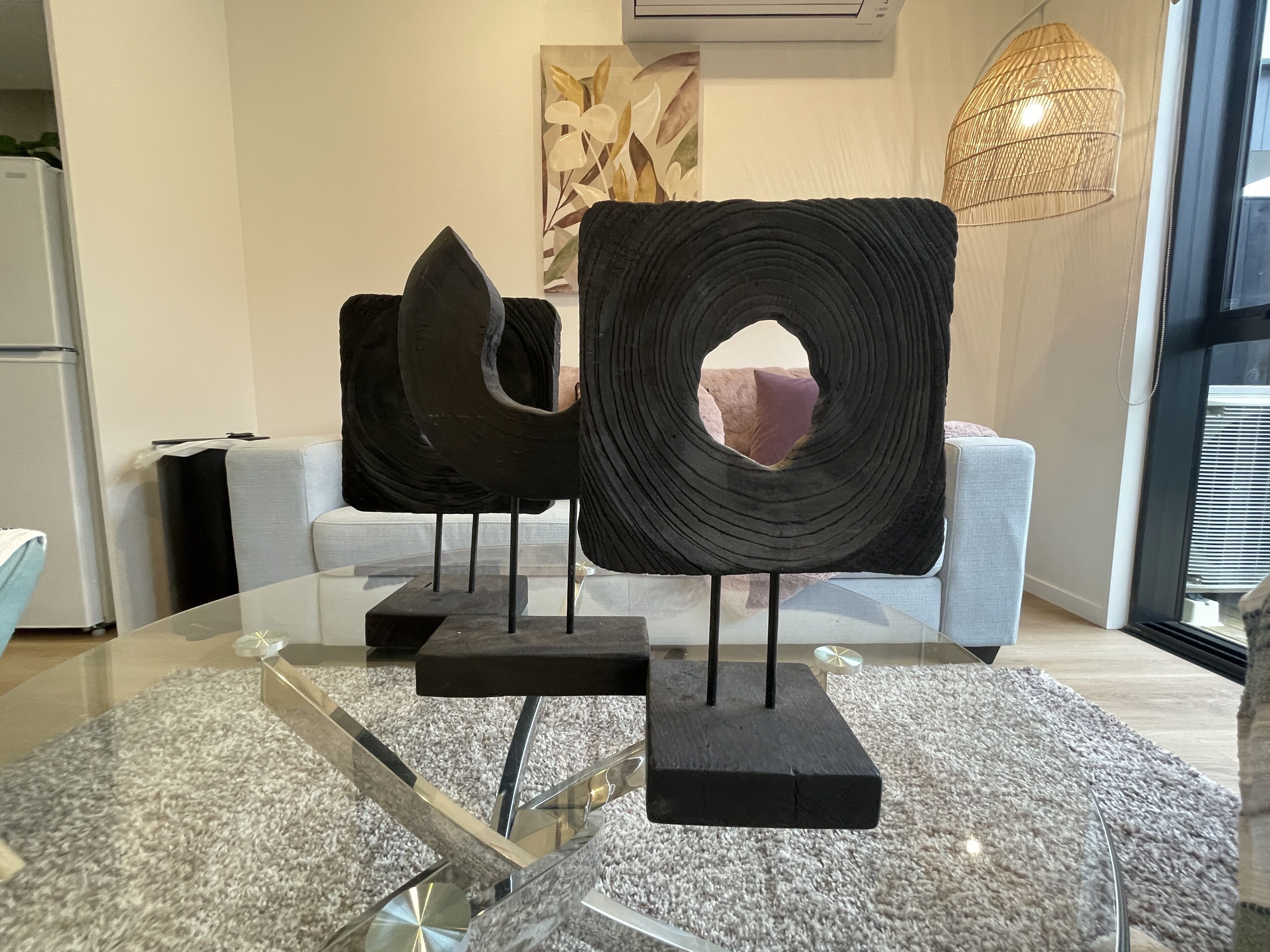 Ein modernes Wohnzimmer mit abstrakten Skulpturen auf Glasfußsochen, einem weißen Sofa und einem großen Fenster mit Jalousien.