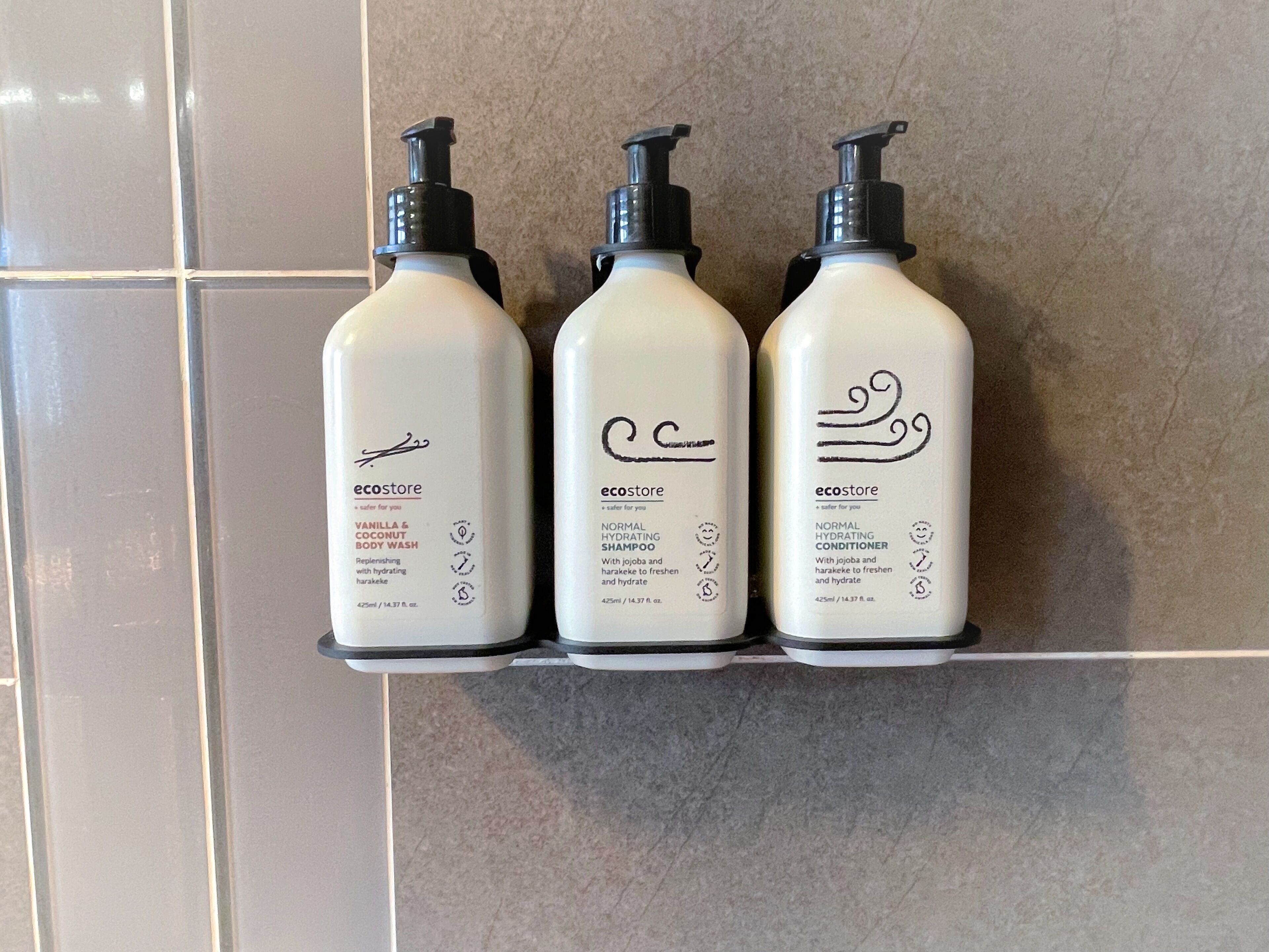 Tres botellas de crema limpiadora y acondicionador para el cuerpo de Ecostore sobre un estante de baldosas.