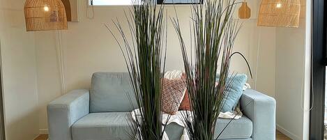 A living room with a light blue sofa, two tall plants in woven pots, a white rug, and a window with blinds.
