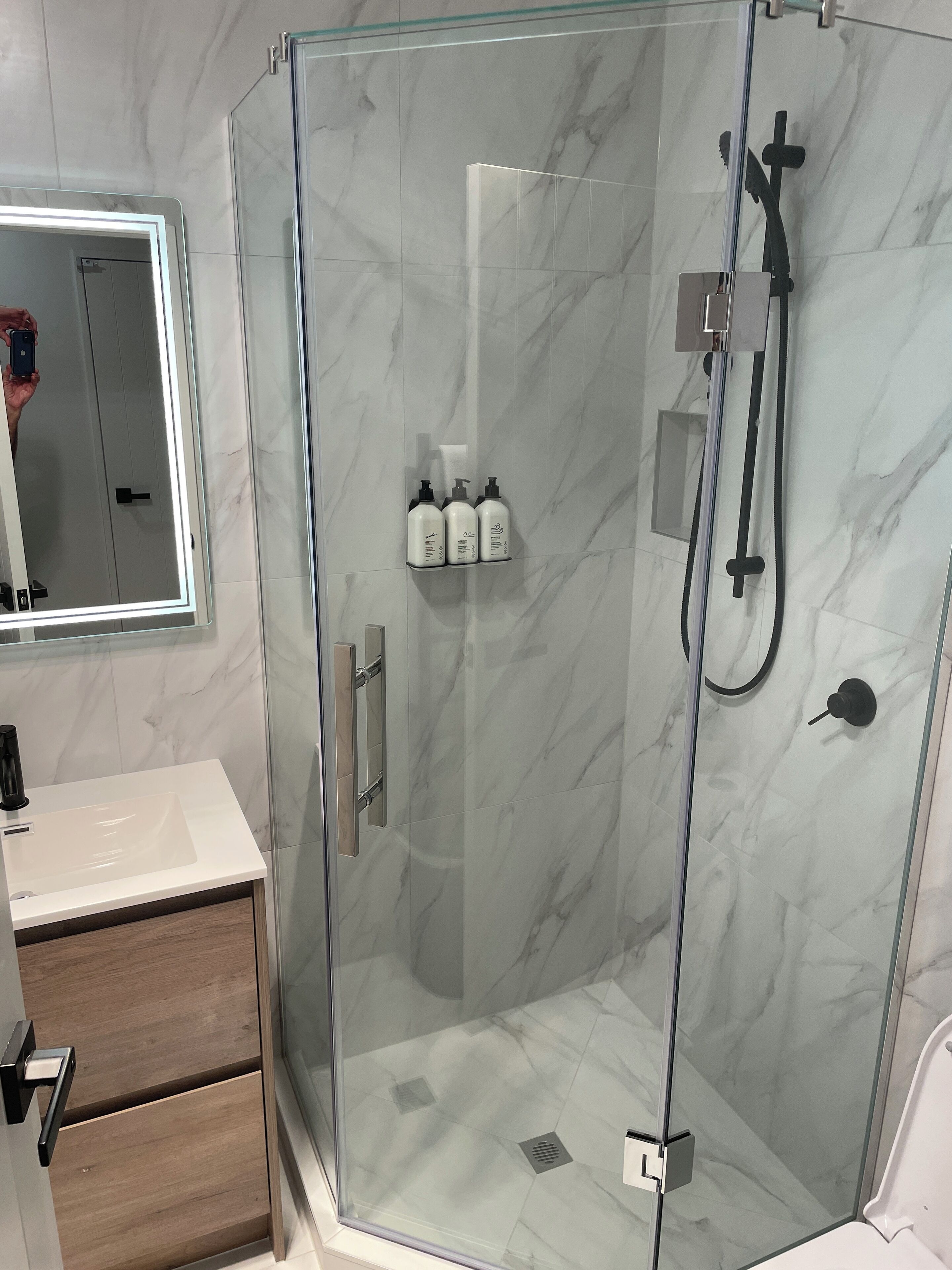 A modern bathroom with a glass-enclosed shower, marble walls, a vanity with a mirror, and toiletries on the wall.