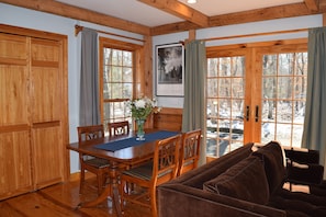 A wooden dining area with a table and chairs, a framed picture on the wall, and a view of the outdoors through large windows.