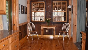 Interior rústico de cabana com paredes de madeira, uma mesa de jantar de madeira, duas cadeiras brancas e uma janela com vista para árvores.