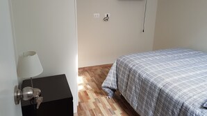 Ein Schlafzimmer mit Bett, einem an der Wand befestigten Fernseher, einem Nachttisch mit Lampe und einer Steckdose.