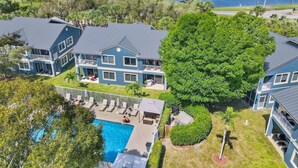 Une grande maison bleue avec une piscine, entourée d’une végétation luxuriante et bordée d’un lac.