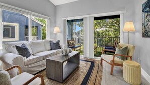 Un salon moderne avec un canapé, une table basse et des fauteuils, de grandes fenêtres et un balcon aménagé avec du mobilier d’extérieur.