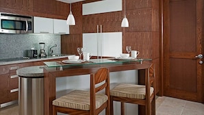 A modern kitchen with wooden cabinetry, a built-in microwave, a glass-top island with two chairs, and pendant lighting.