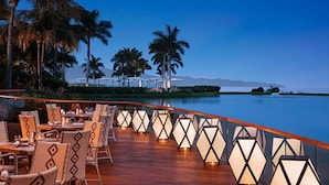 A deck with tables and chairs overlooking a calm body of water, palm trees, and distant mountains.