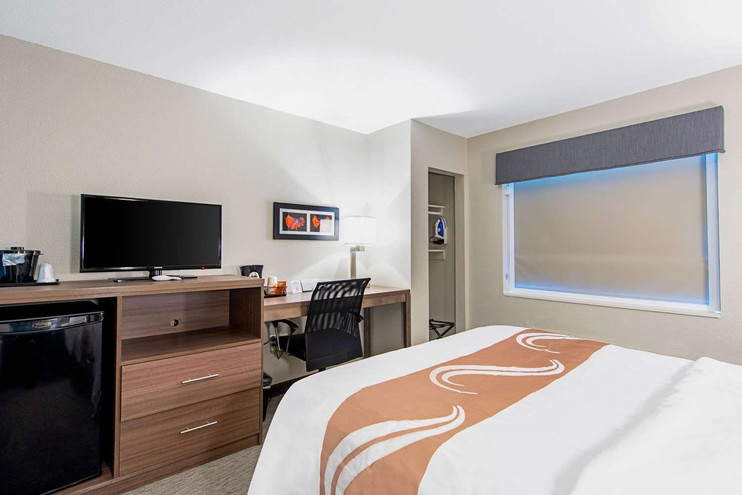 A hotel room with a bed, a desk with a computer, a television, and a window with blinds.