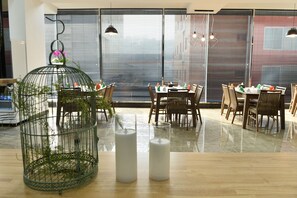 Un coin repas avec des tables et des chaises en bois, une grande fenêtre avec des stores et une cage à oiseaux verte posée sur la table.