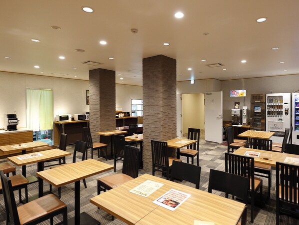 Un restaurante de hotel con varias mesas y sillas, una máquina de café, un refrigerador y una pantalla de televisión.