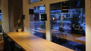 Una larga mesa de madera con sillas negras, ventanas grandes con persianas y vistas a la calle.