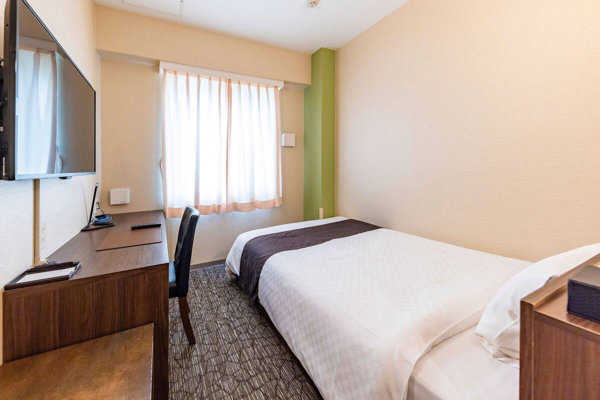 Une chambre d’hôtel comprenant un lit, un bureau avec une chaise, une télévision et une fenêtre avec des rideaux.