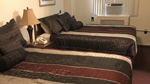 A hotel room with two beds, a bedside table with a lamp, a telephone, and a window with blinds. - The Mountaineer Hotel (Williamson)