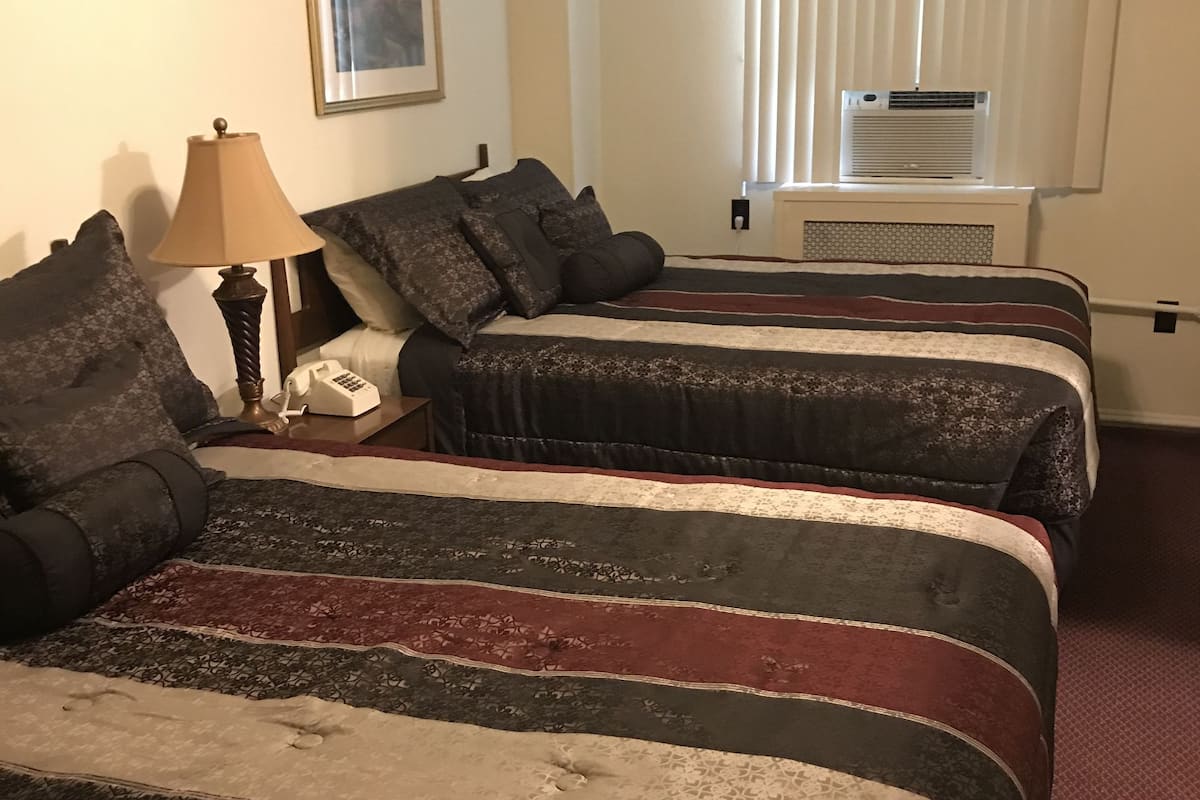 A hotel room with two beds, a bedside table with a lamp, a telephone, and a window with blinds.