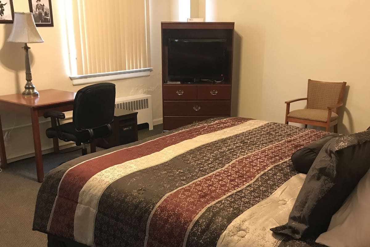 A hotel room with a bed, a desk with a lamp, a chair, a TV on a dresser, and a window with blinds.