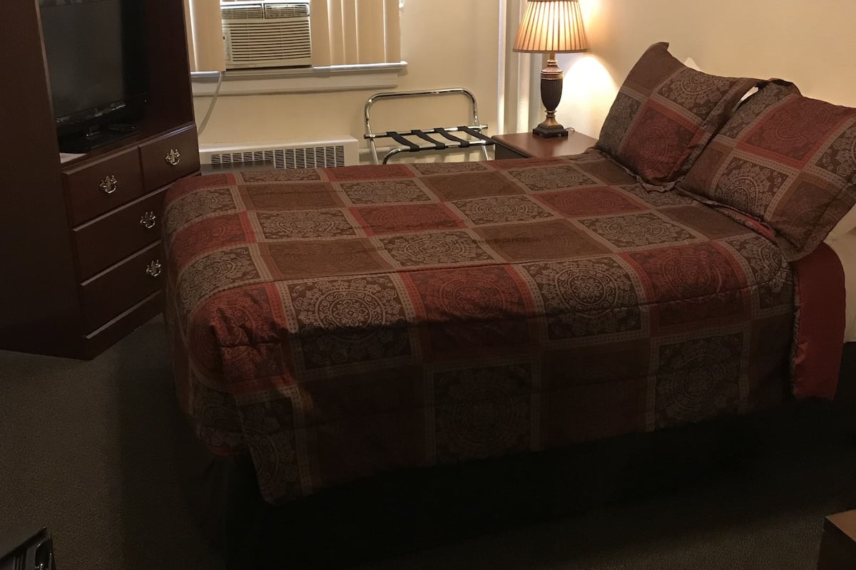 A hotel room with a bed, a TV, a lamp, and a window with blinds.