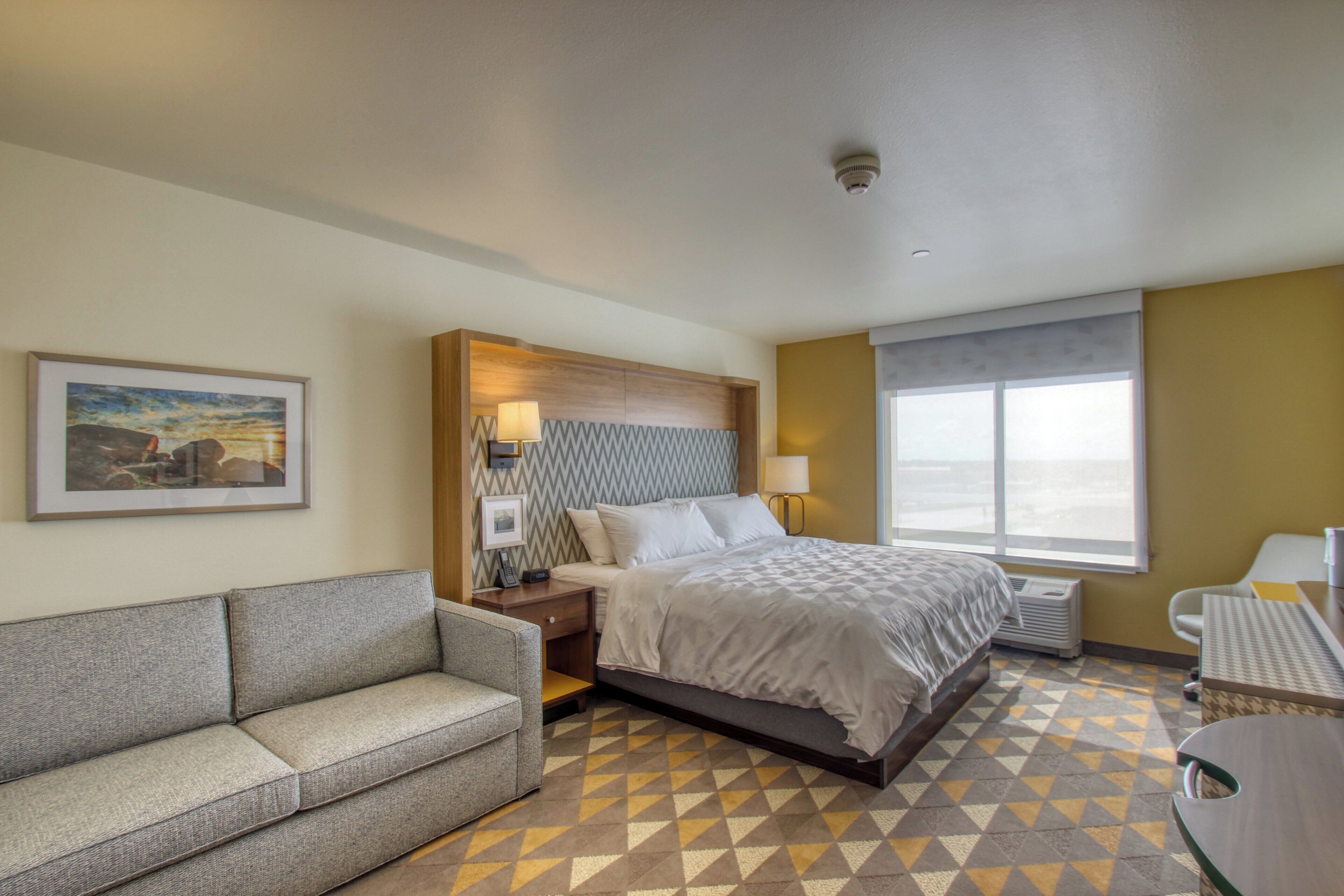 A hotel room with a bed, a sofa, a desk, a window, and a painting on the wall.