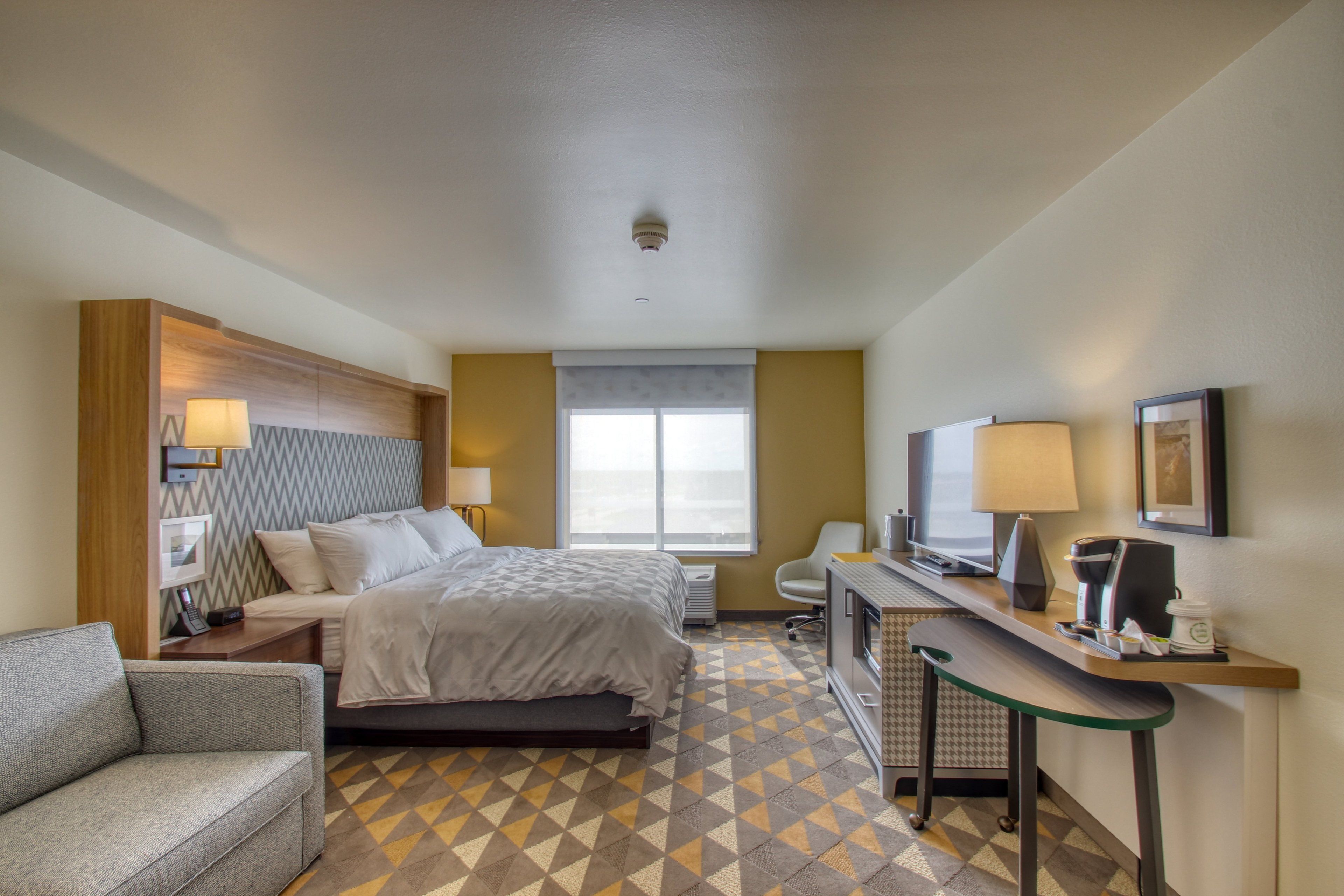 A hotel room with a bed, a desk with a computer, a chair, a sofa, a window, and a wall-mounted TV.