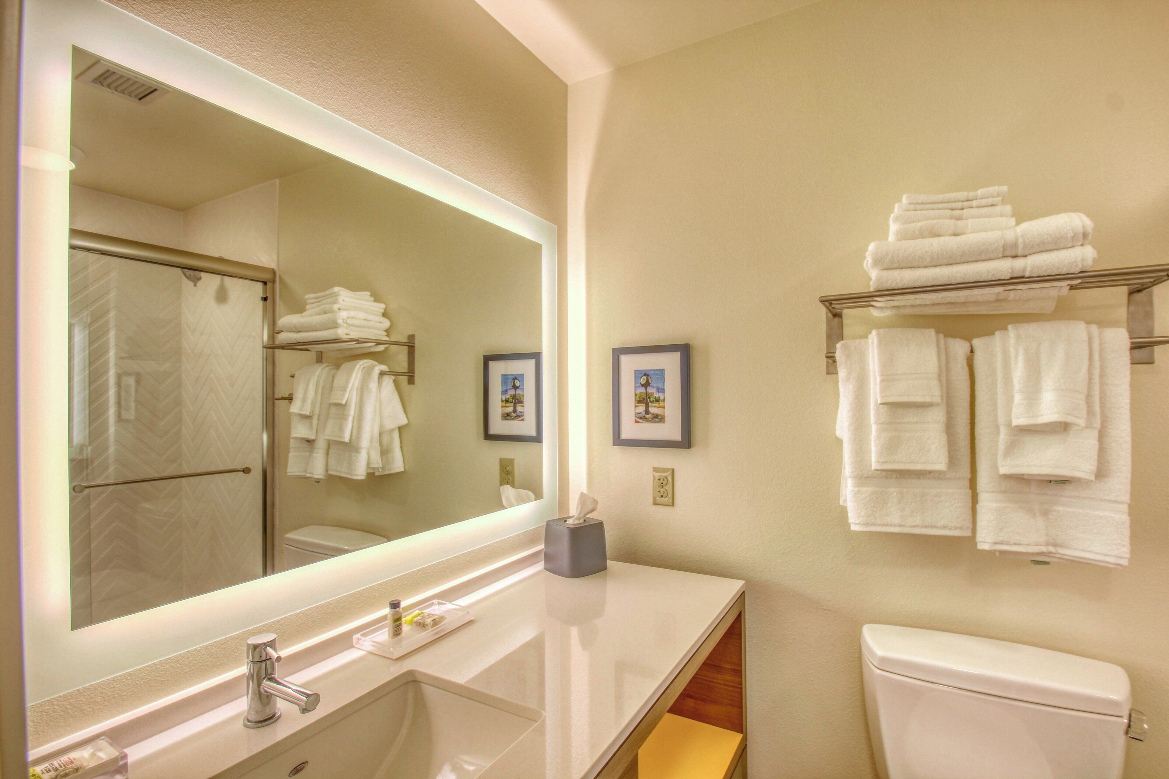 Un baño de hotel con un espejo grande, un lavamanos, un inodoro y un perchero con toallas dobladas.