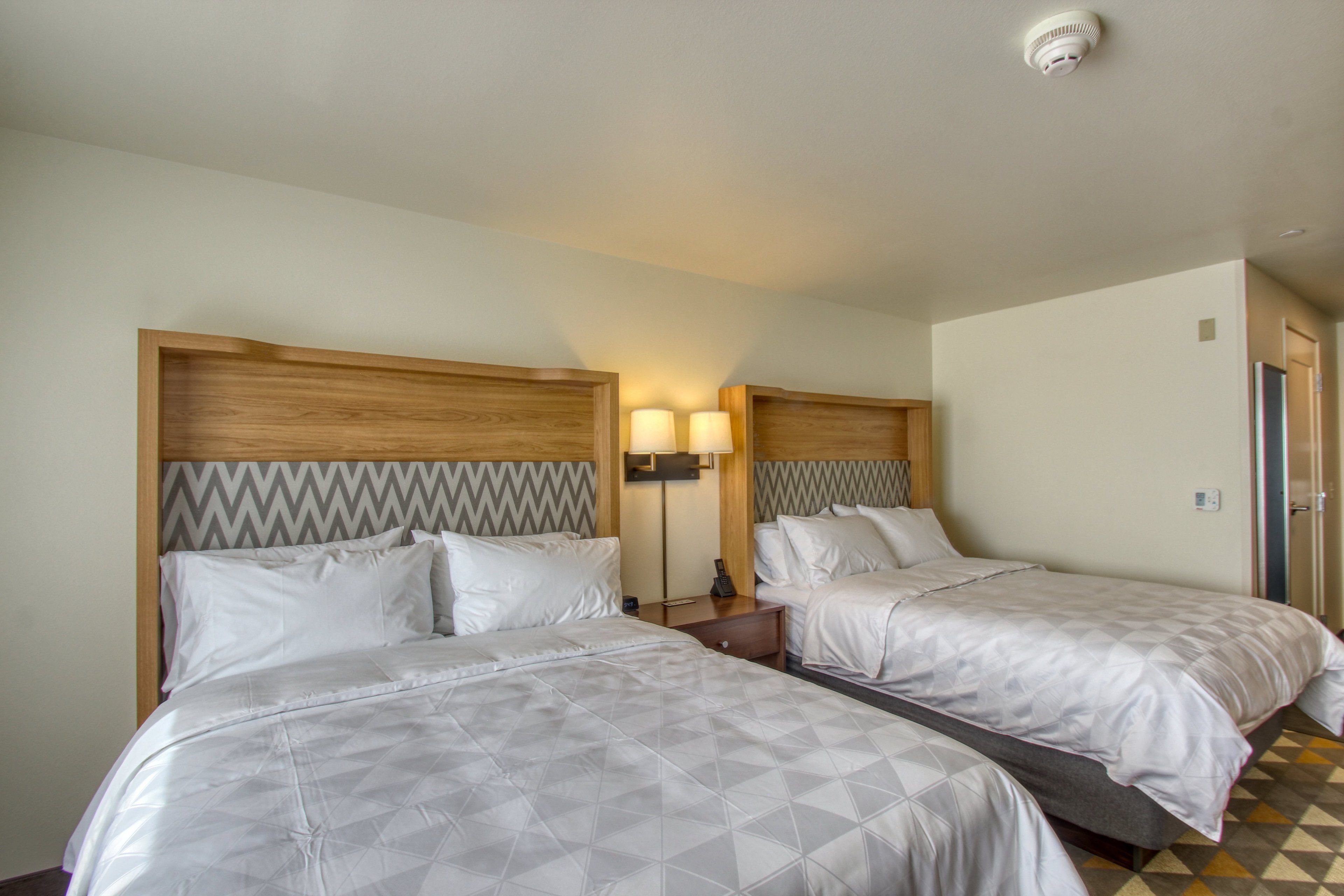 A hotel room with two beds, a wooden headboard with a zigzag pattern, a bedside table with a lamp, and a patterned floor.