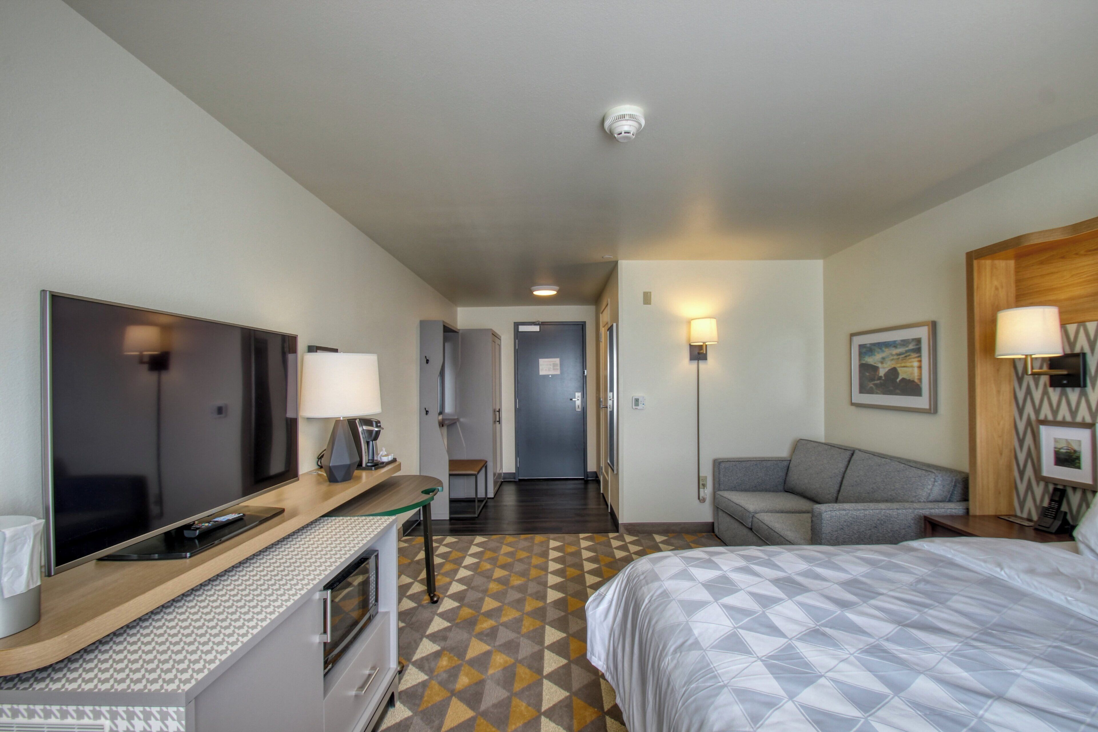 A hotel room with a bed, a TV, a desk with a lamp, a sofa, and a hallway leading to a door.