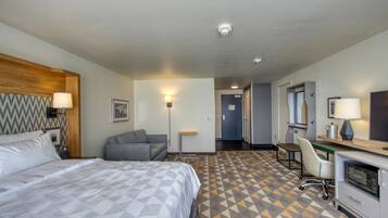 Habitación de hotel con cama, escritorio, silla, microondas y una mesita de noche con lámpara.