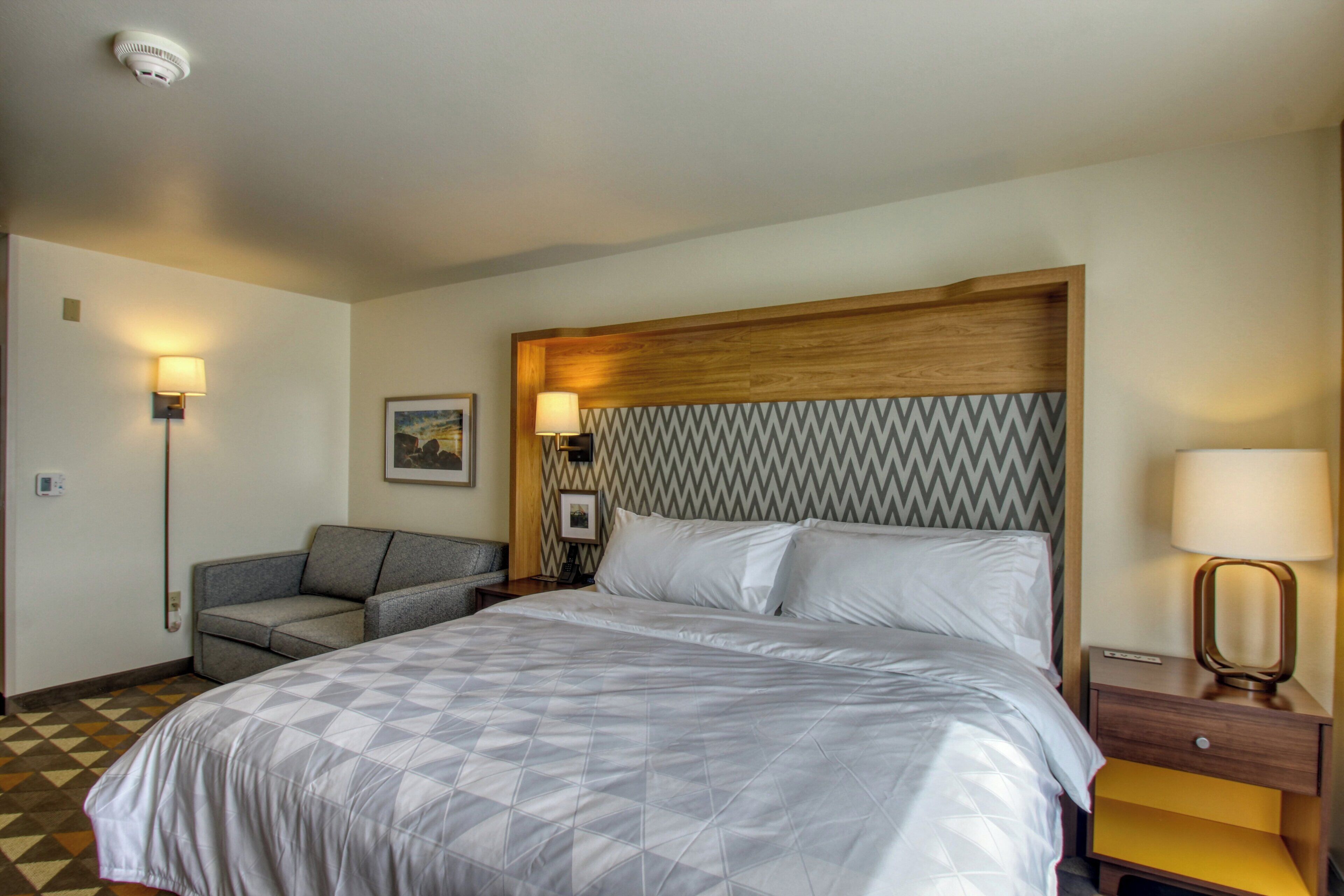 A hotel room with a large bed, a sofa, a nightstand with a lamp, and a wall-mounted picture.
