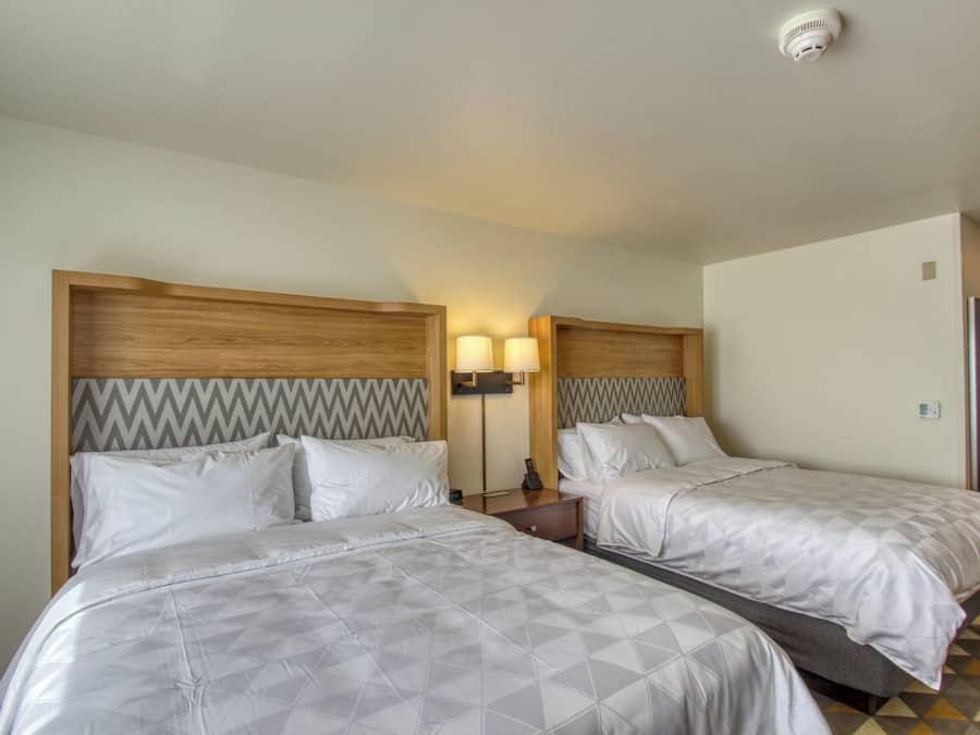 Habitación de hotel con dos camas, cabecero de madera con diseño en zigzag, mesita de noche con lámpara y suelo de baldosas.