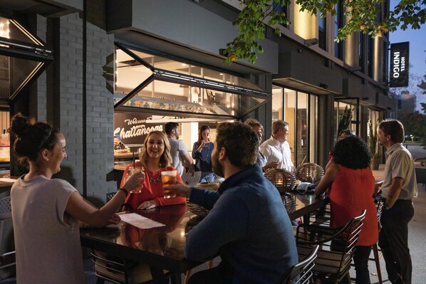 Un espace extérieur aménagé avec des tables et des chaises, un espace clos en verre pour le comptoir de service de restauration, et une enseigne indiquant que l’établissement est un hôtel.