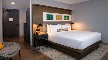 A modern hotel room with a large bed, bedside tables, a desk, a chair, and artwork on the wall.