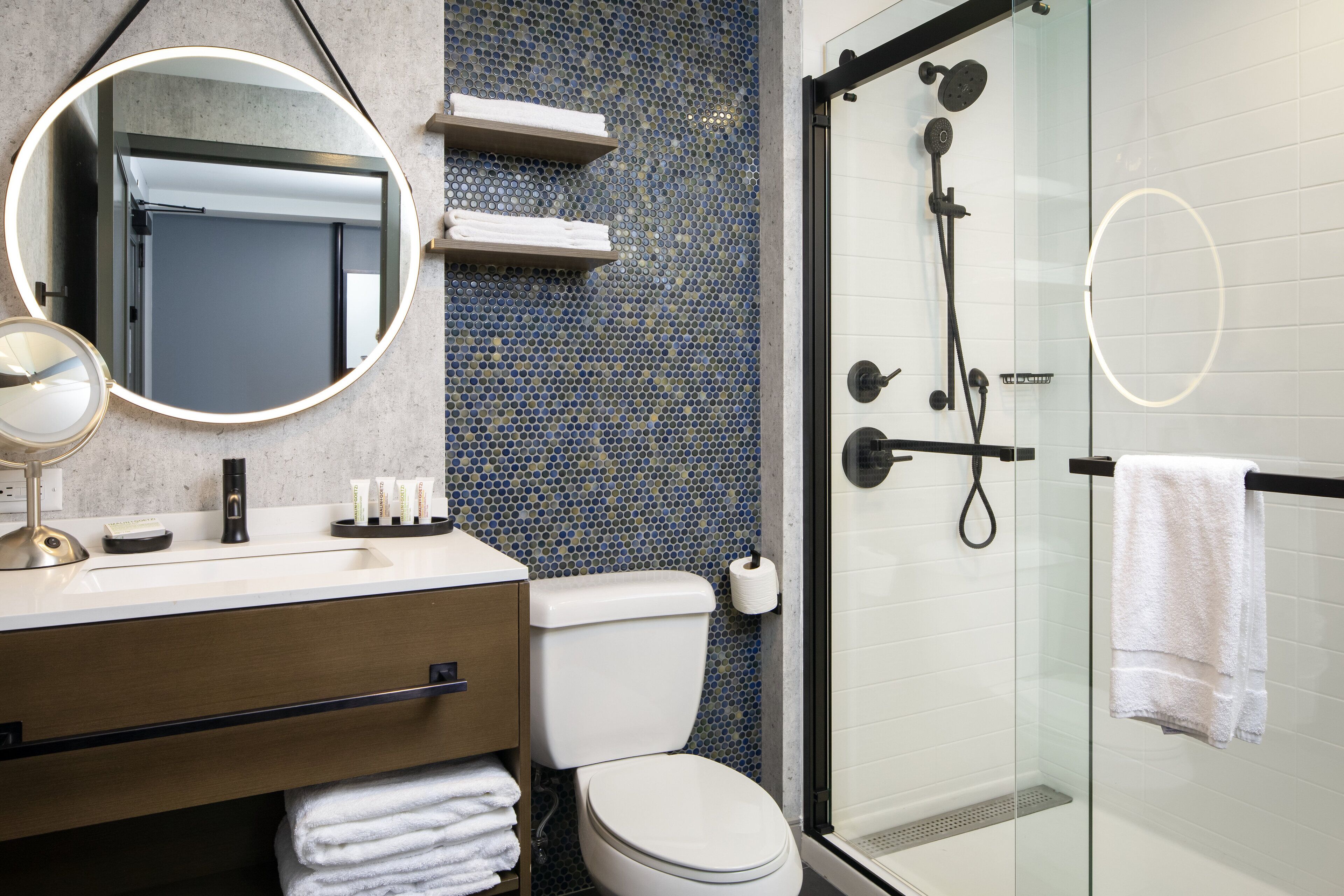 Un bagno moderno con cabina doccia in vetro, uno specchio rotondo, un water e un lavabo con porta asciugamani.