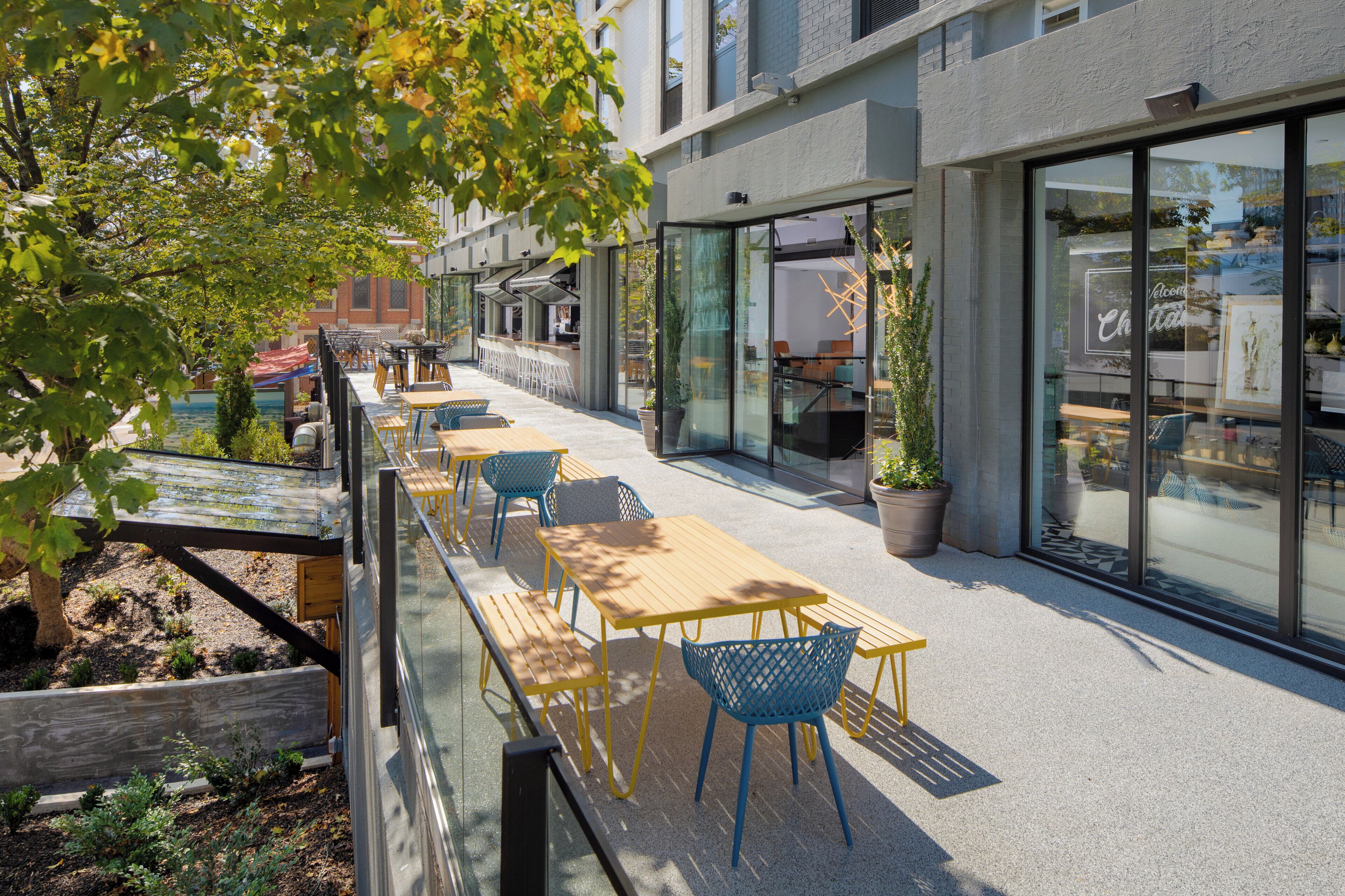 An outdoor seating area with tables and chairs, a glass wall, and potted plants.
