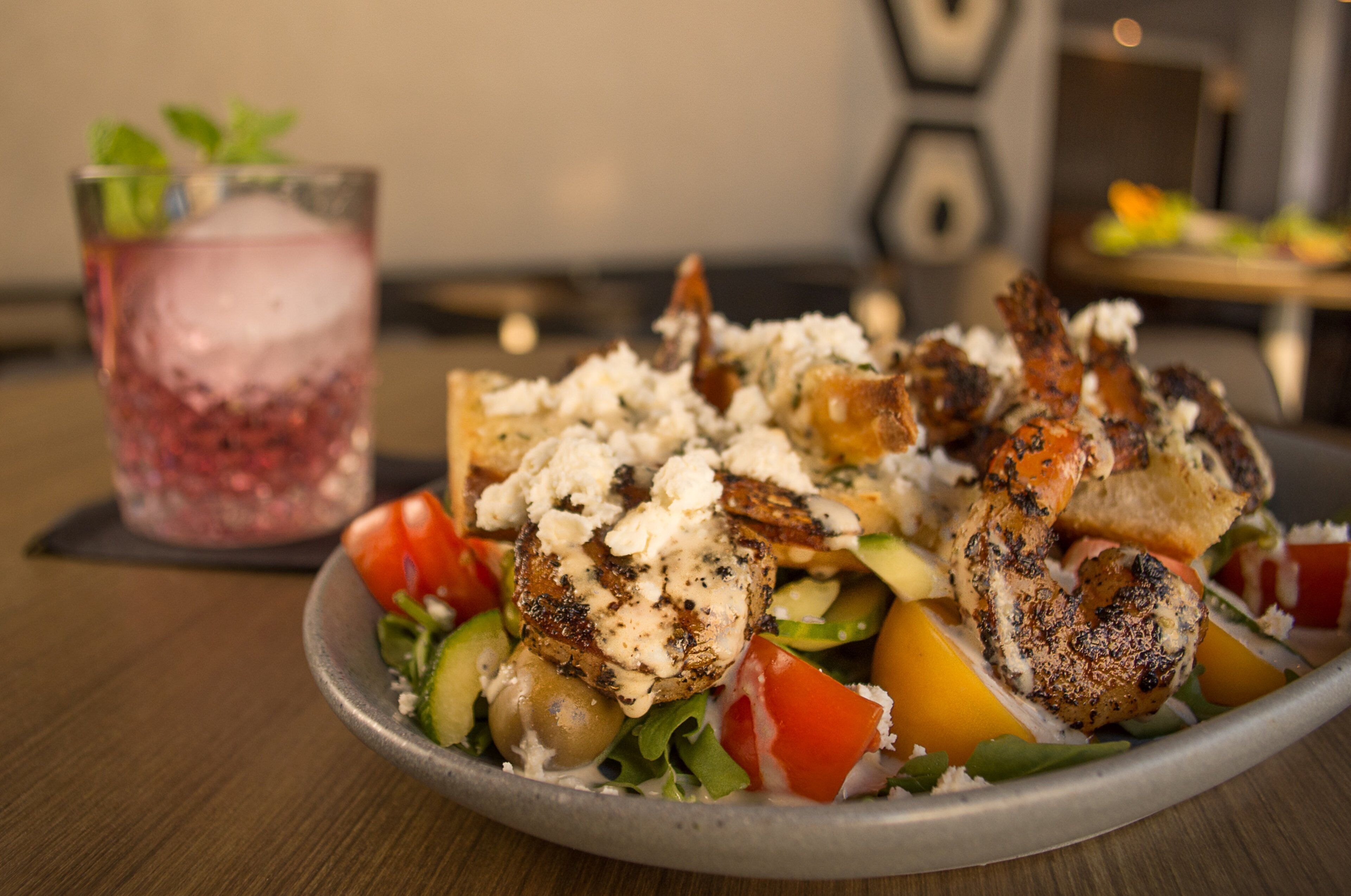 Un plato de pollo a la parrilla, queso feta y verduras sobre una mesa, con un vaso de bebida helada al fondo.