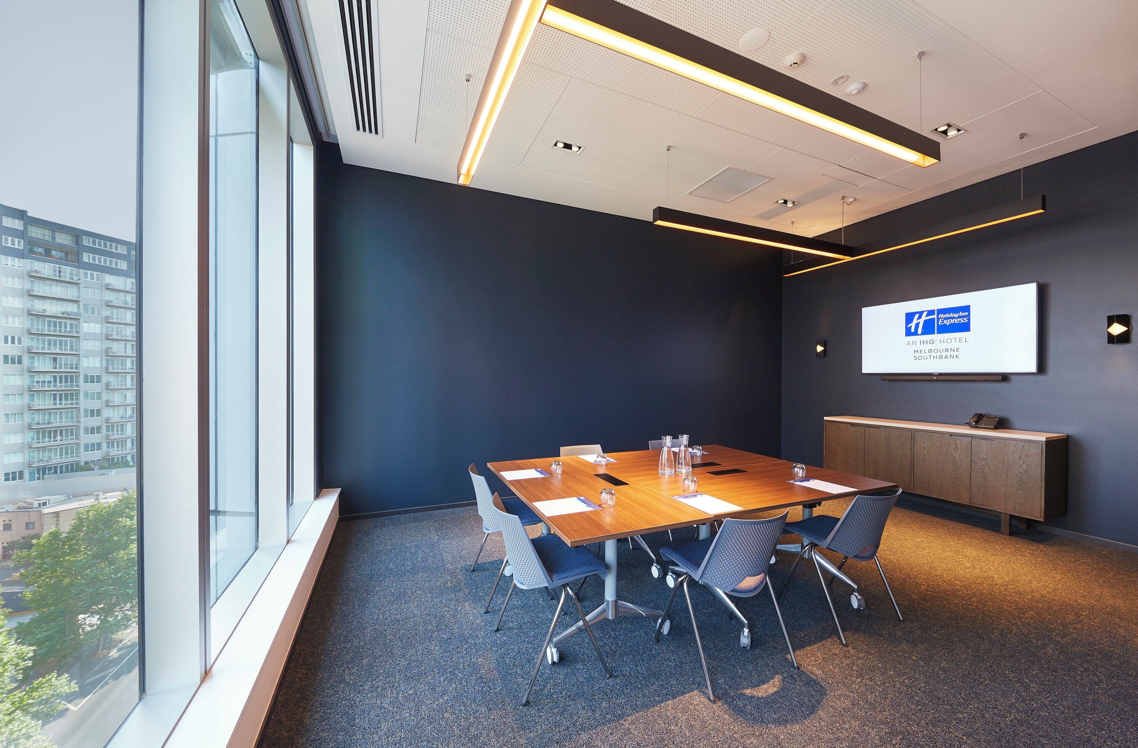 A conference room with a large table, chairs, a projector screen, and a view of a building through large windows.